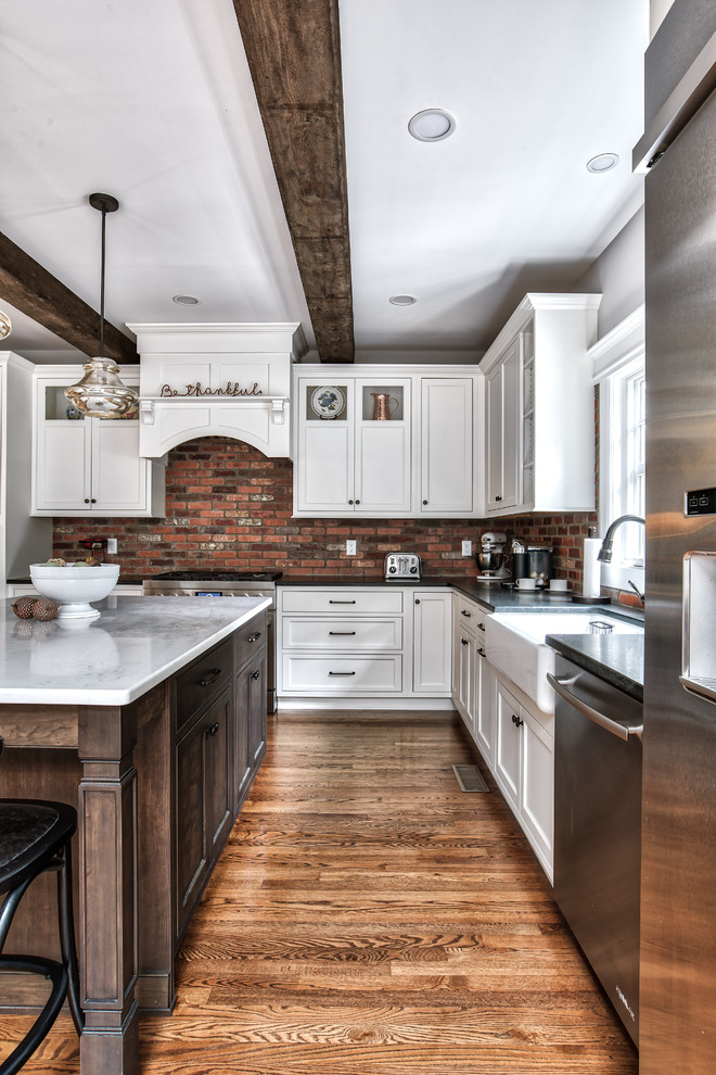 Large country l-shaped eat-in kitchen in New York with a farmhouse sink, beaded inset cabinets, white cabinets, granite benchtops, brick splashback, stainless steel appliances, with island, brown floor, black benchtop and painted wood floors.