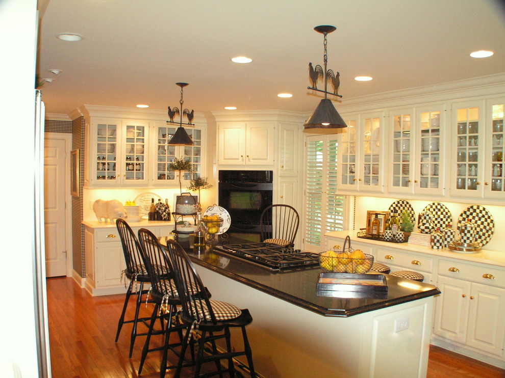 Photo of a mid-sized country single-wall eat-in kitchen in Other with glass-front cabinets, white cabinets, solid surface benchtops, white splashback, black appliances, medium hardwood floors and with island.