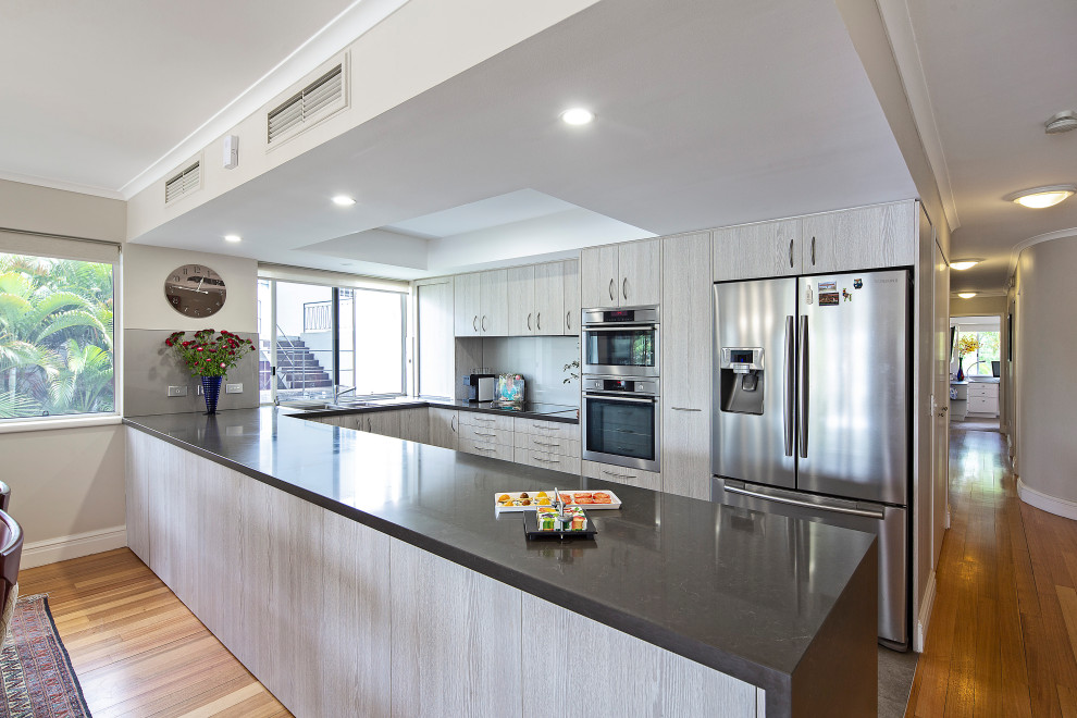This is an example of a mid-sized transitional u-shaped kitchen pantry in Brisbane with an undermount sink, flat-panel cabinets, light wood cabinets, quartz benchtops, white splashback, glass sheet splashback, stainless steel appliances, medium hardwood floors, a peninsula, brown floor and black benchtop.