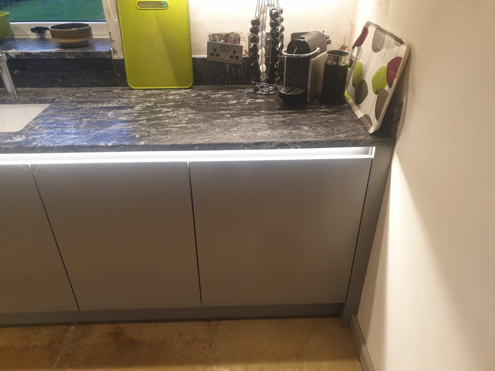Mid-sized contemporary l-shaped eat-in kitchen in West Midlands with flat-panel cabinets, granite benchtops, black splashback, engineered quartz splashback, stainless steel appliances, black benchtop, a drop-in sink, black cabinets, travertine floors, a peninsula, beige floor and vaulted.