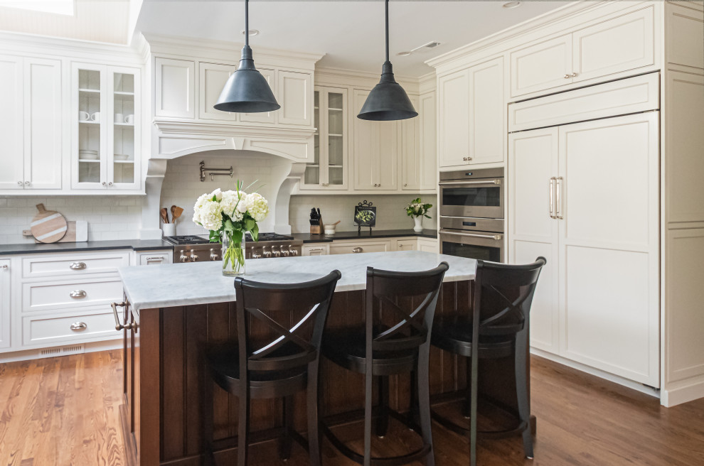 Design ideas for a large traditional u-shaped open plan kitchen in Philadelphia with a farmhouse sink, beaded inset cabinets, white cabinets, granite benchtops, white splashback, subway tile splashback, panelled appliances, medium hardwood floors, with island, brown floor and black benchtop.