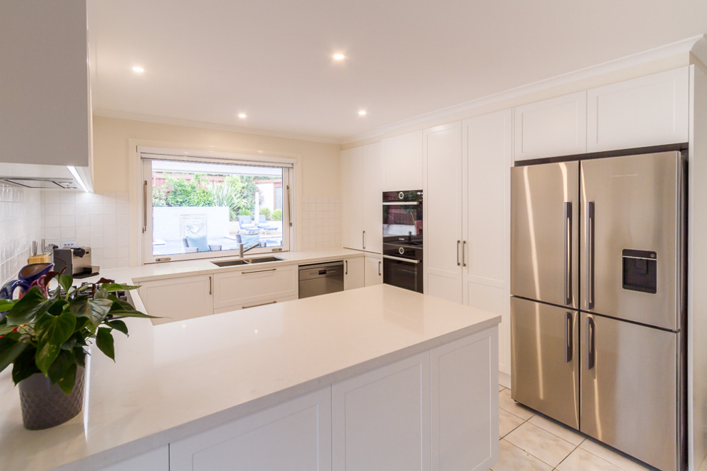 Photo of a mid-sized traditional u-shaped eat-in kitchen in Melbourne with a double-bowl sink, shaker cabinets, white cabinets, quartz benchtops, white splashback, porcelain splashback, stainless steel appliances, porcelain floors, a peninsula, beige floor and white benchtop.