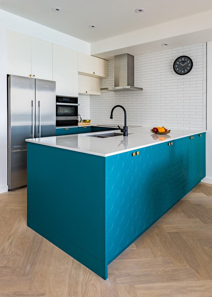 This is an example of a small scandinavian u-shaped eat-in kitchen in Toronto with an undermount sink, flat-panel cabinets, blue cabinets, quartz benchtops, white splashback, porcelain splashback, stainless steel appliances, light hardwood floors, a peninsula and white benchtop.