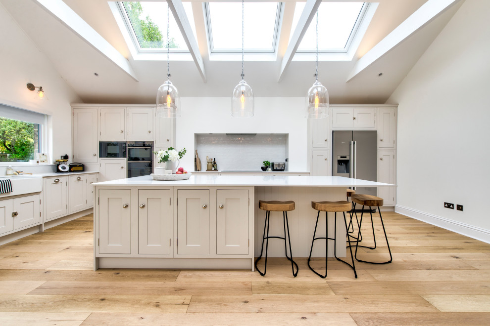 Photo of a large transitional l-shaped kitchen in Glasgow with a farmhouse sink, shaker cabinets, beige cabinets, white splashback, glass tile splashback, stainless steel appliances, light hardwood floors, with island, beige floor and white benchtop.