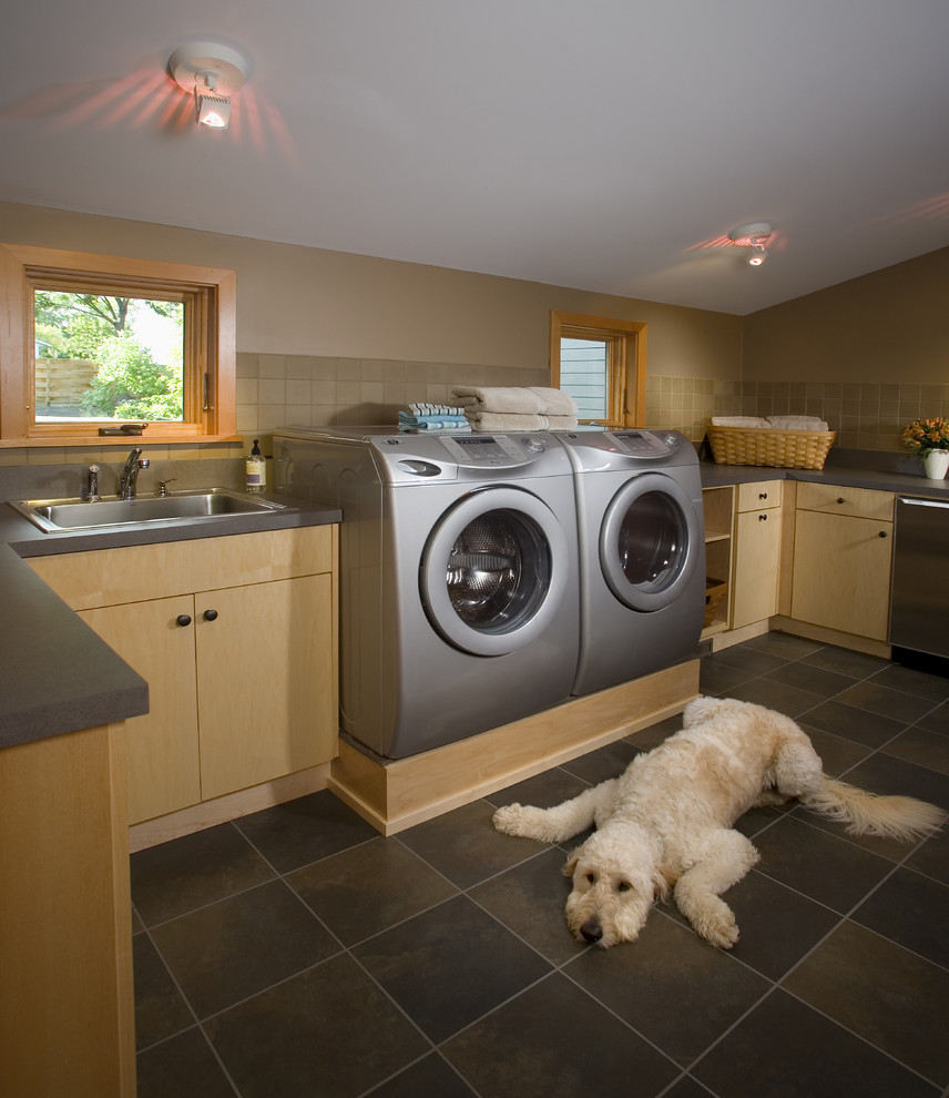 This is an example of a large contemporary u-shaped dedicated laundry room in Minneapolis with light wood cabinets, a drop-in sink, flat-panel cabinets, solid surface benchtops, beige walls, travertine floors, a side-by-side washer and dryer, grey floor and grey benchtop.