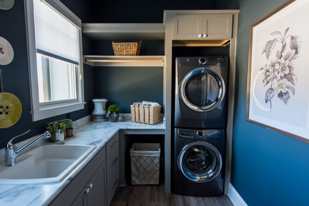 Photo of a mid-sized transitional l-shaped dedicated laundry room in Indianapolis with a drop-in sink, shaker cabinets, grey cabinets, blue walls, medium hardwood floors, a stacked washer and dryer, brown floor and grey benchtop.