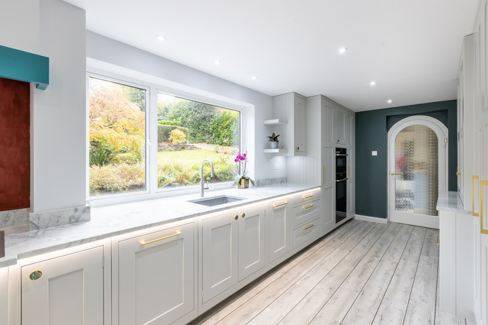Large contemporary grey and teal open plan kitchen in Sussex with shaker cabinets, grey cabinets, quartz worktops, black appliances, an island, grey floors and grey worktops.