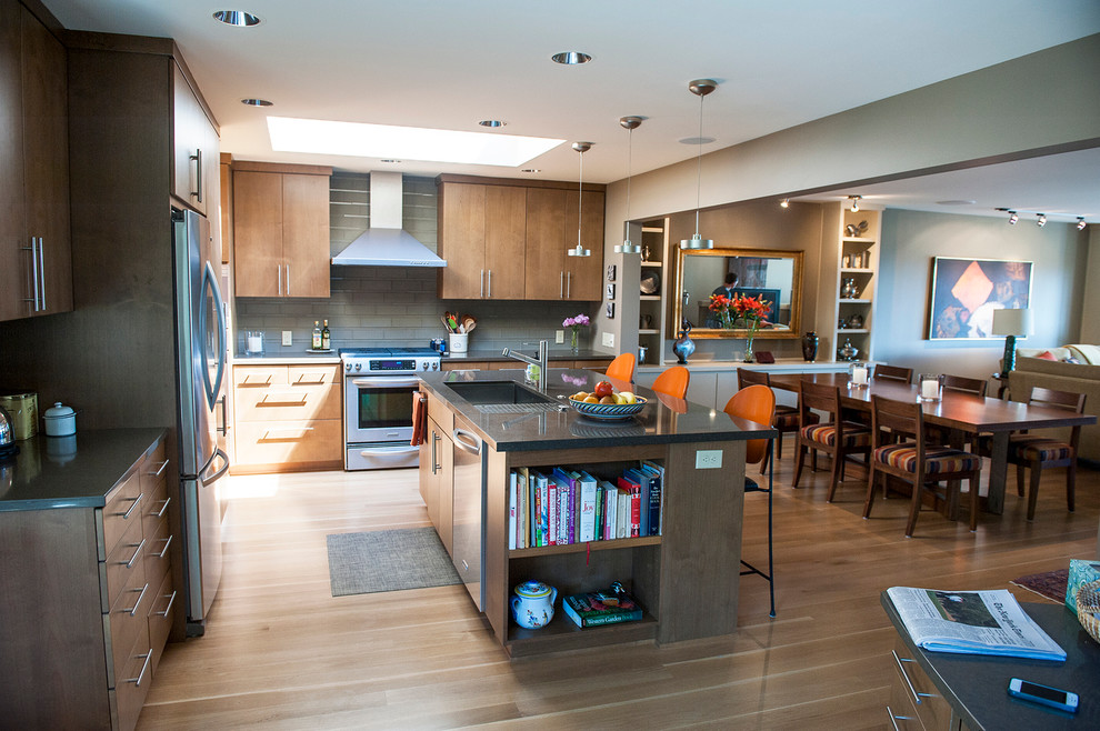 This is an example of a mid-sized contemporary open plan kitchen in Portland with a triple-bowl sink, flat-panel cabinets, medium wood cabinets, quartz benchtops, grey splashback, subway tile splashback, stainless steel appliances, light hardwood floors and with island.