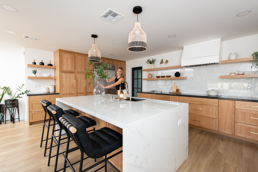 Photo of a large beach style l-shaped eat-in kitchen in Orange County with a drop-in sink, shaker cabinets, medium wood cabinets, quartzite benchtops, multi-coloured splashback, stone slab splashback, stainless steel appliances, light hardwood floors, with island and white benchtop.
