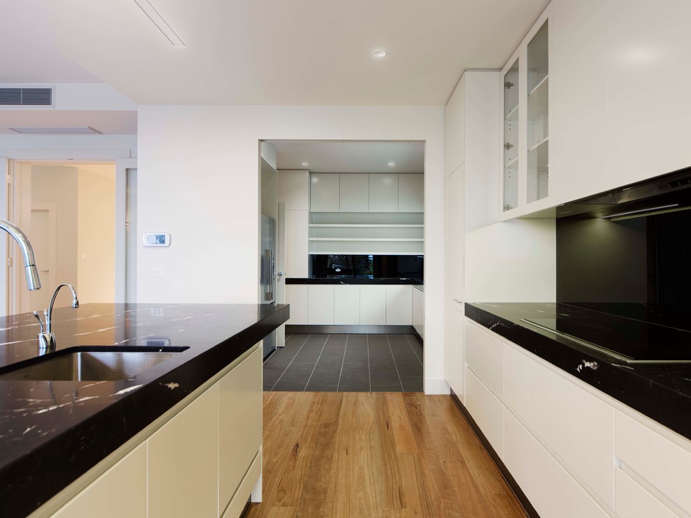 Inspiration for a large contemporary galley open plan kitchen in Melbourne with an undermount sink, flat-panel cabinets, white cabinets, marble benchtops, black splashback, glass sheet splashback, stainless steel appliances, light hardwood floors and multiple islands.