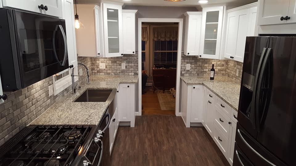 Mid-sized country galley separate kitchen in Boston with a farmhouse sink, shaker cabinets, white cabinets, quartz benchtops, brown splashback, stone tile splashback, black appliances, dark hardwood floors, no island, brown floor and beige benchtop.