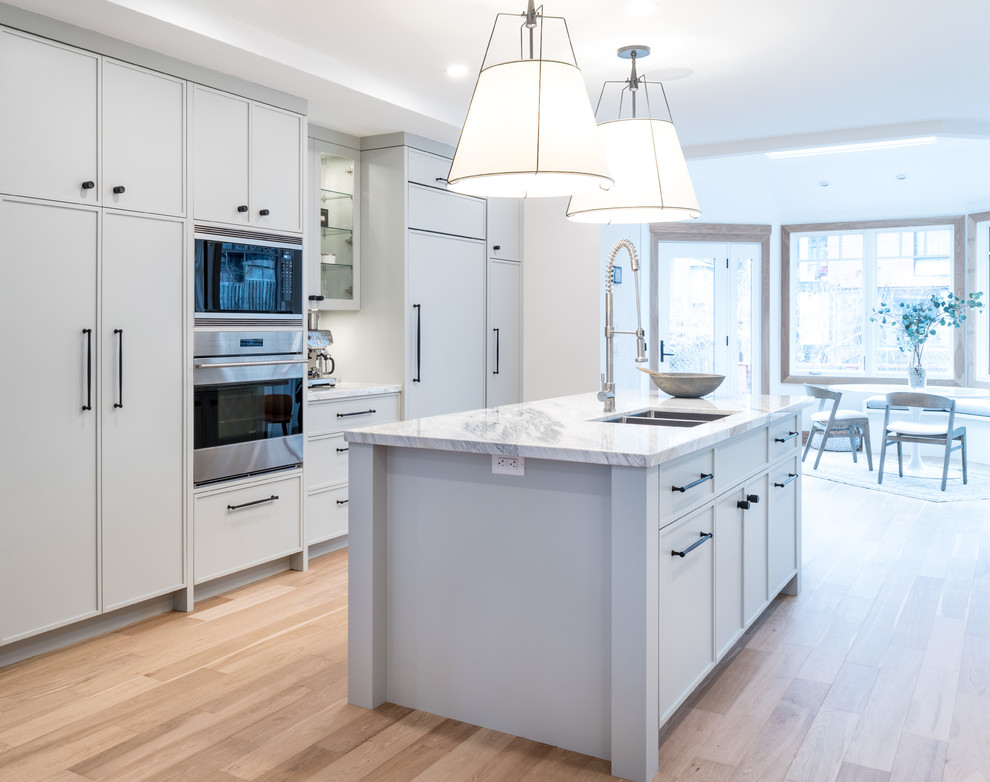 This is an example of a large beach style galley open plan kitchen in Toronto with an undermount sink, shaker cabinets, grey cabinets, marble benchtops, white splashback, marble splashback, stainless steel appliances, light hardwood floors, with island, beige floor and white benchtop.