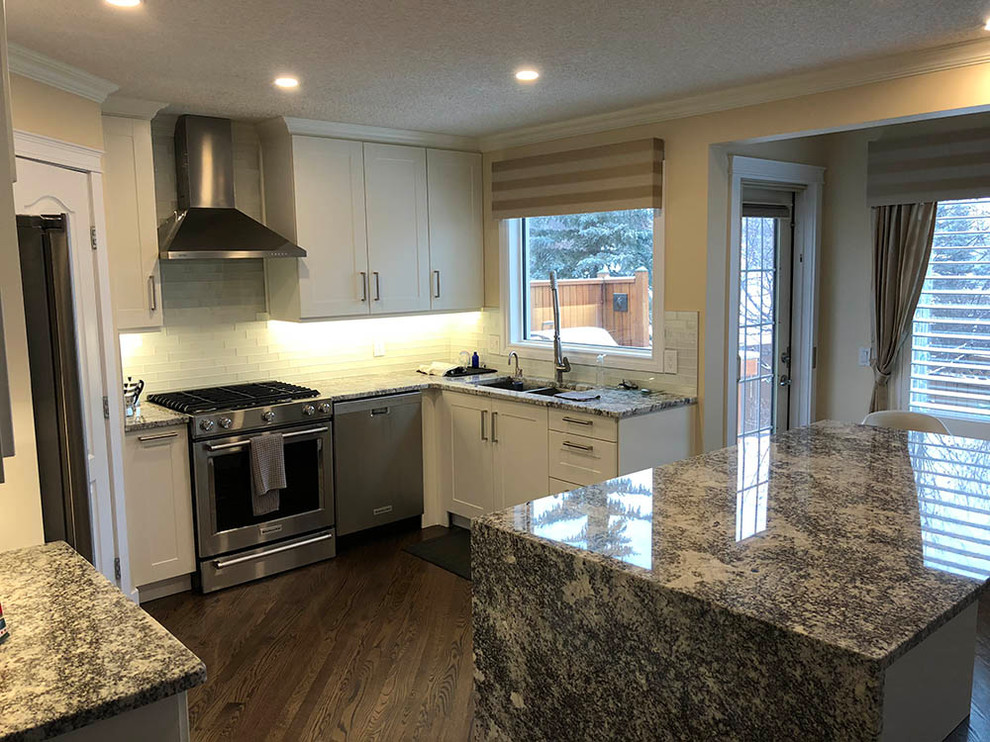 Inspiration for a mid-sized transitional u-shaped medium tone wood floor and brown floor enclosed kitchen remodel in Calgary with a double-bowl sink, beige cabinets, granite countertops, white backsplash, subway tile backsplash, stainless steel appliances, multicolored countertops, shaker cabinets and an island