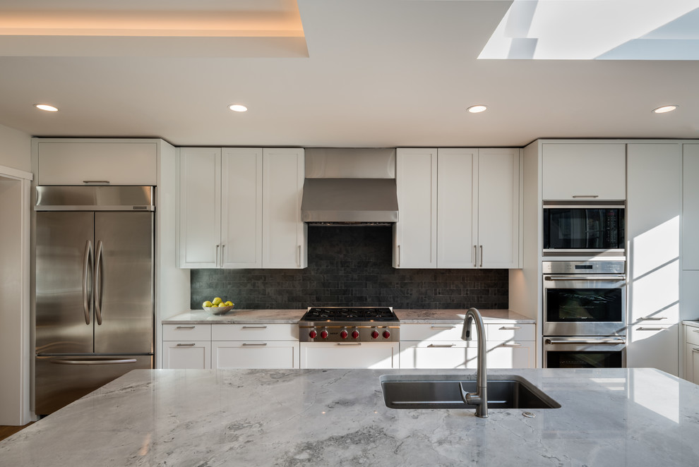 Photo of a large contemporary u-shaped separate kitchen in DC Metro with a drop-in sink, recessed-panel cabinets, white cabinets, black splashback, slate splashback, stainless steel appliances, light hardwood floors, with island, brown floor and quartzite benchtops.