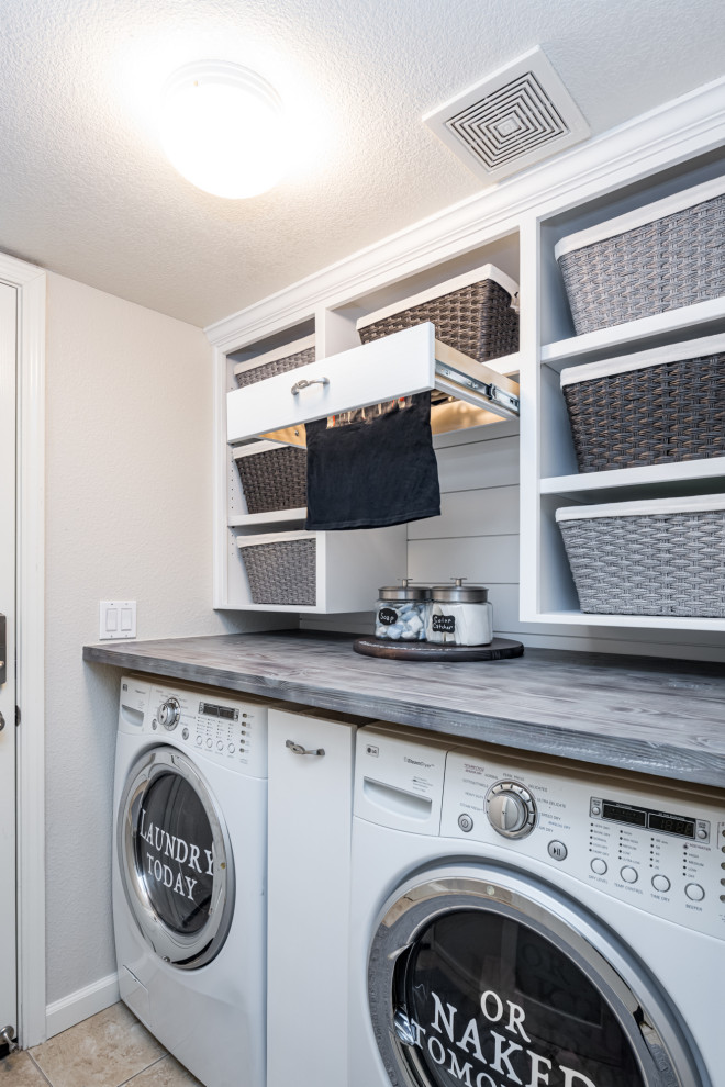 Photo of a small country single-wall utility room in Sacramento with open cabinets, white cabinets, wood benchtops, white walls, porcelain floors, a side-by-side washer and dryer, beige floor and grey benchtop.