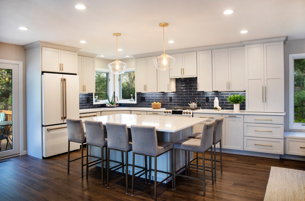 This is an example of a mid-sized transitional l-shaped open plan kitchen in Other with an undermount sink, shaker cabinets, white cabinets, quartz benchtops, blue splashback, subway tile splashback, white appliances, medium hardwood floors, with island, brown floor and white benchtop.