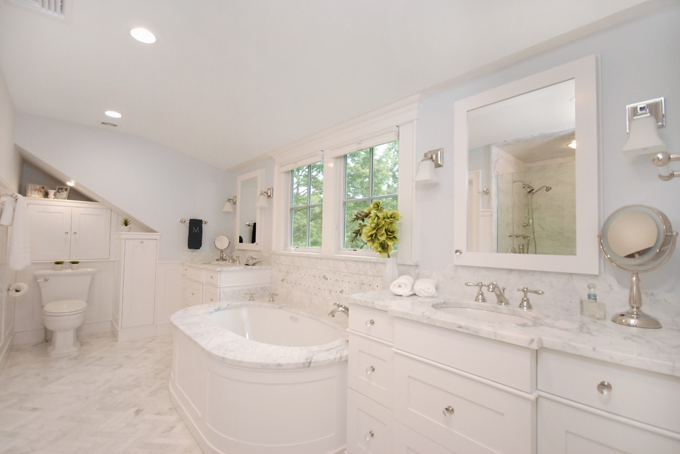 This is an example of a large master bathroom in New York with shaker cabinets, white cabinets, an undermount tub, a one-piece toilet, white tile, white walls, marble floors and an undermount sink.