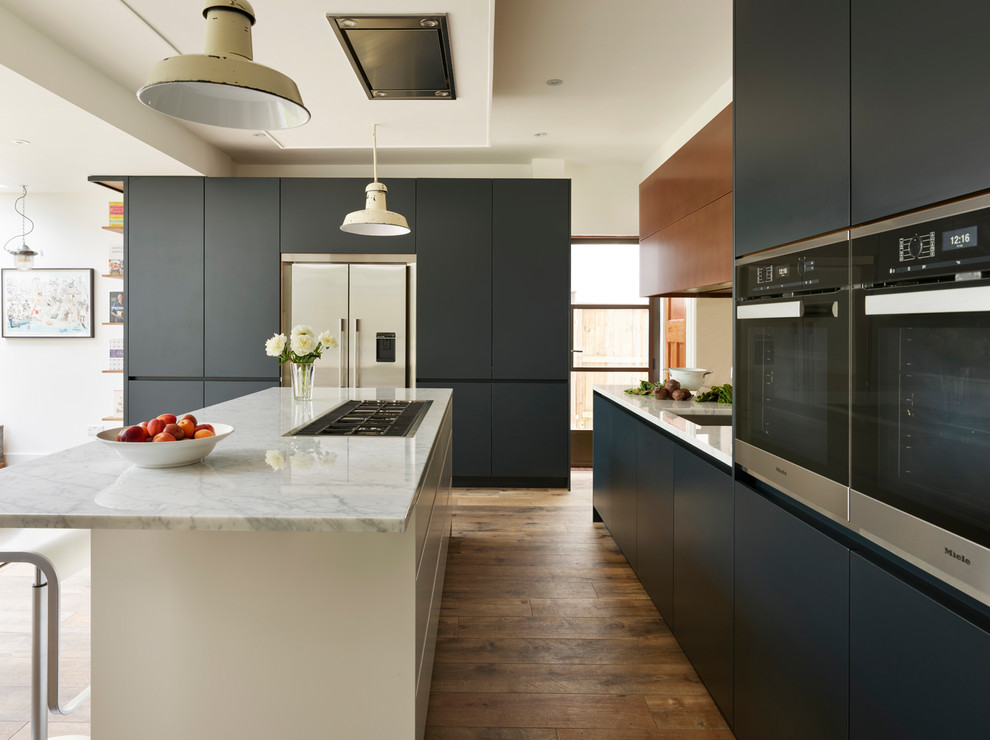 Inspiration for a large contemporary l-shaped eat-in kitchen in London with an integrated sink, blue cabinets, marble benchtops, metallic splashback, mirror splashback, light hardwood floors, with island, brown floor and white benchtop.