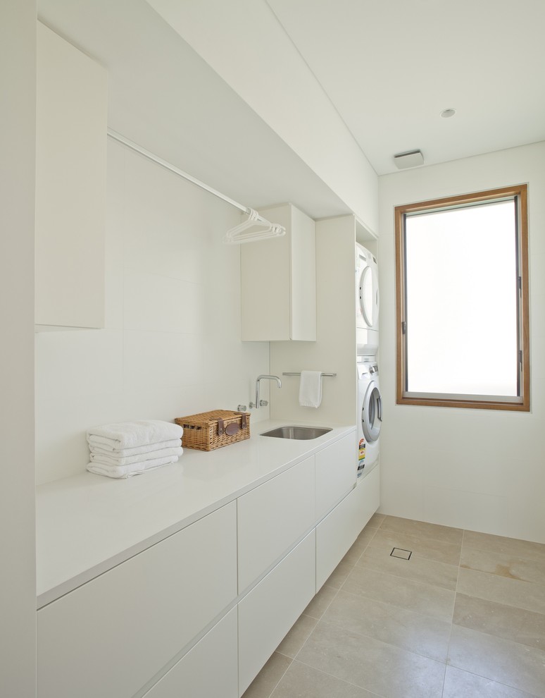 Large contemporary single-wall dedicated laundry room in Sydney with a single-bowl sink, flat-panel cabinets, white cabinets, terrazzo benchtops, white walls, limestone floors, a stacked washer and dryer, beige floor and white benchtop.