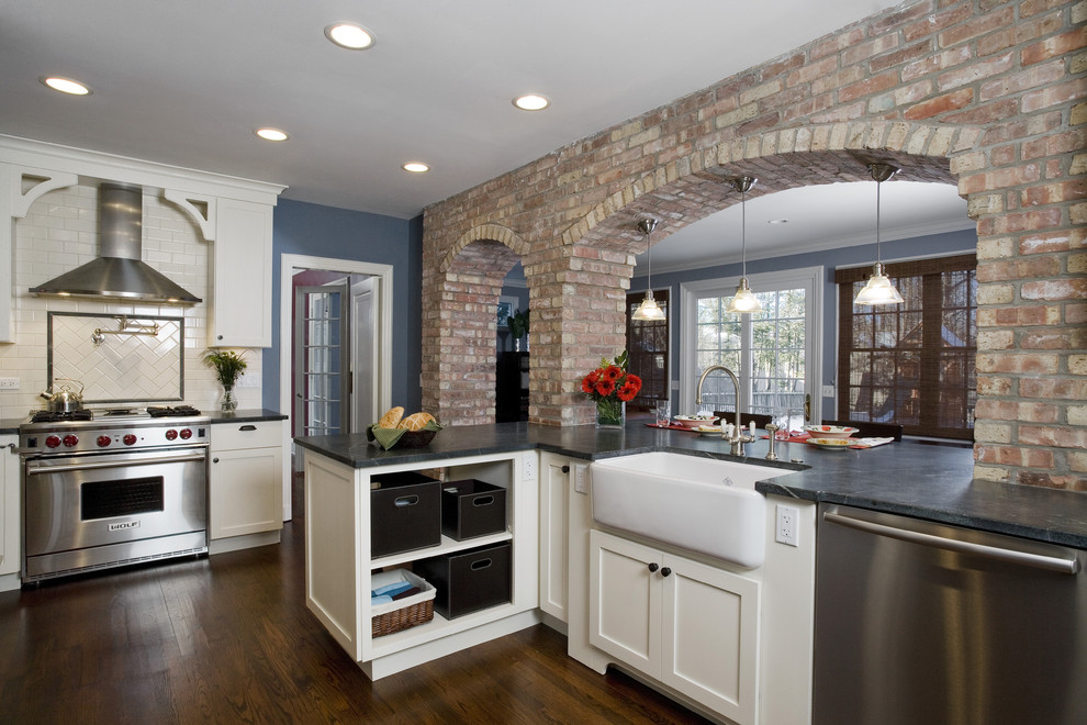 Design ideas for a mid-sized traditional u-shaped eat-in kitchen in Chicago with stainless steel appliances, subway tile splashback, a farmhouse sink, soapstone benchtops, shaker cabinets, white cabinets, white splashback, medium hardwood floors, brown floor and black benchtop.
