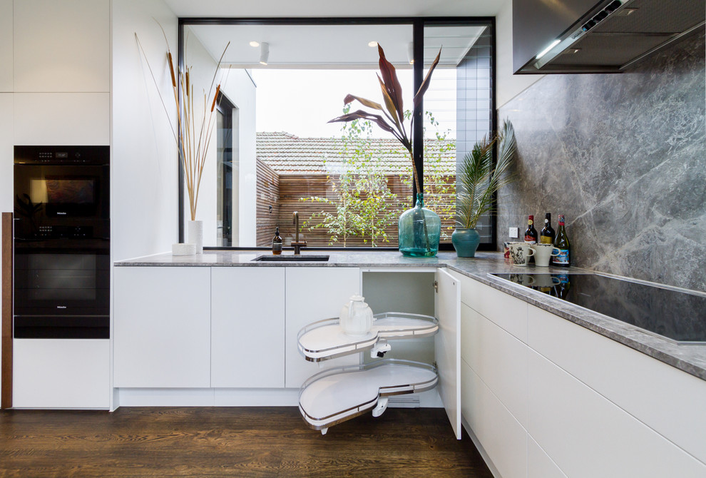 This is an example of a large modern l-shaped open plan kitchen in Melbourne with an undermount sink, flat-panel cabinets, white cabinets, grey splashback, marble splashback, black appliances, medium hardwood floors, with island, brown floor, grey benchtop and limestone benchtops.