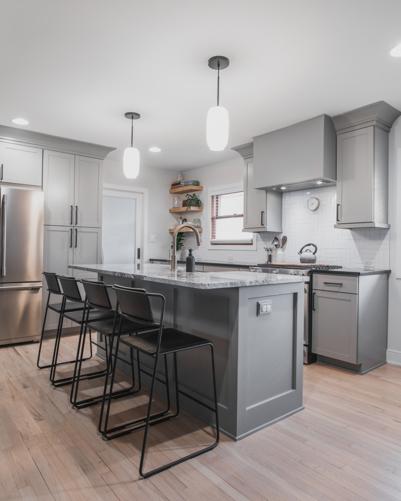 Inspiration for a mid-sized modern l-shaped eat-in kitchen in Chicago with a farmhouse sink, shaker cabinets, grey cabinets, granite benchtops, white splashback, ceramic splashback, stainless steel appliances, light hardwood floors, with island and multi-coloured benchtop.