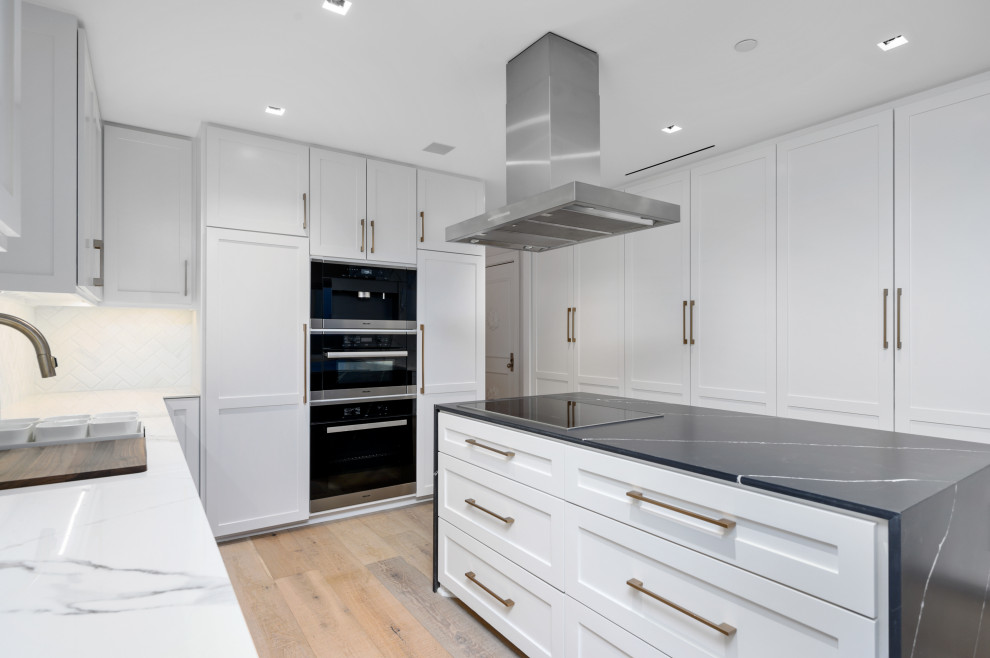 This is an example of a large modern galley eat-in kitchen in Houston with an undermount sink, raised-panel cabinets, white cabinets, solid surface benchtops, white splashback, marble splashback, stainless steel appliances, light hardwood floors, with island, brown floor and black benchtop.