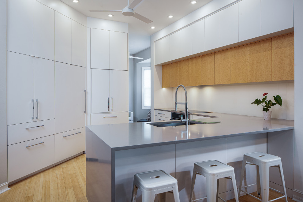 Mid-sized contemporary u-shaped open plan kitchen in Miami with an undermount sink, flat-panel cabinets, white cabinets, quartz benchtops, white splashback, glass sheet splashback, panelled appliances, bamboo floors, a peninsula, yellow floor and grey benchtop.