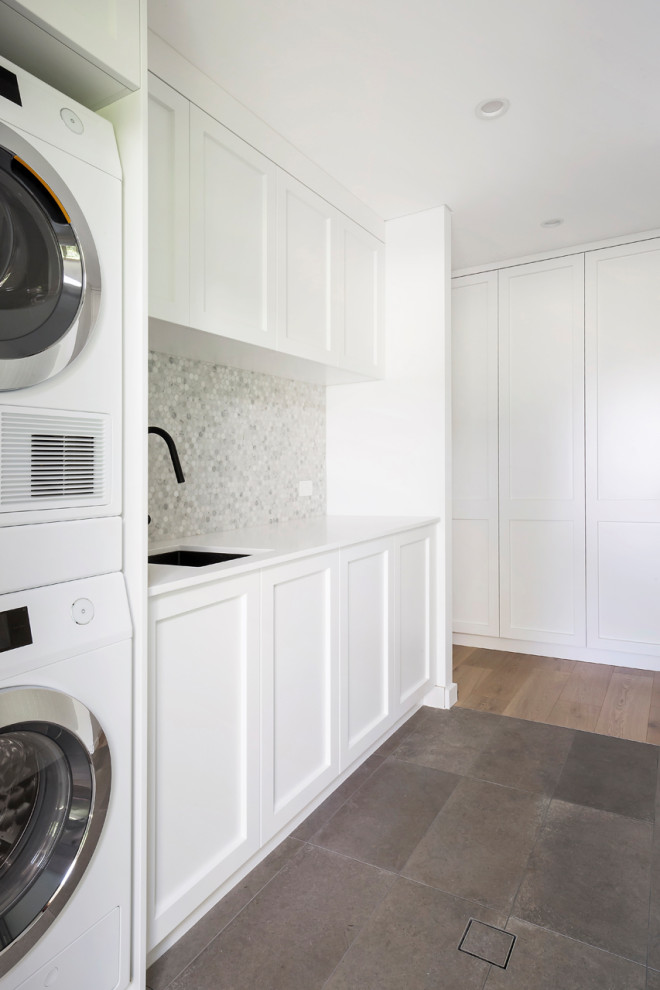 Inspiration for a mid-sized beach style galley dedicated laundry room in Sydney with an undermount sink, shaker cabinets, white cabinets, quartz benchtops, grey splashback, mosaic tile splashback, white walls, porcelain floors, a stacked washer and dryer, grey floor, white benchtop, coffered and panelled walls.