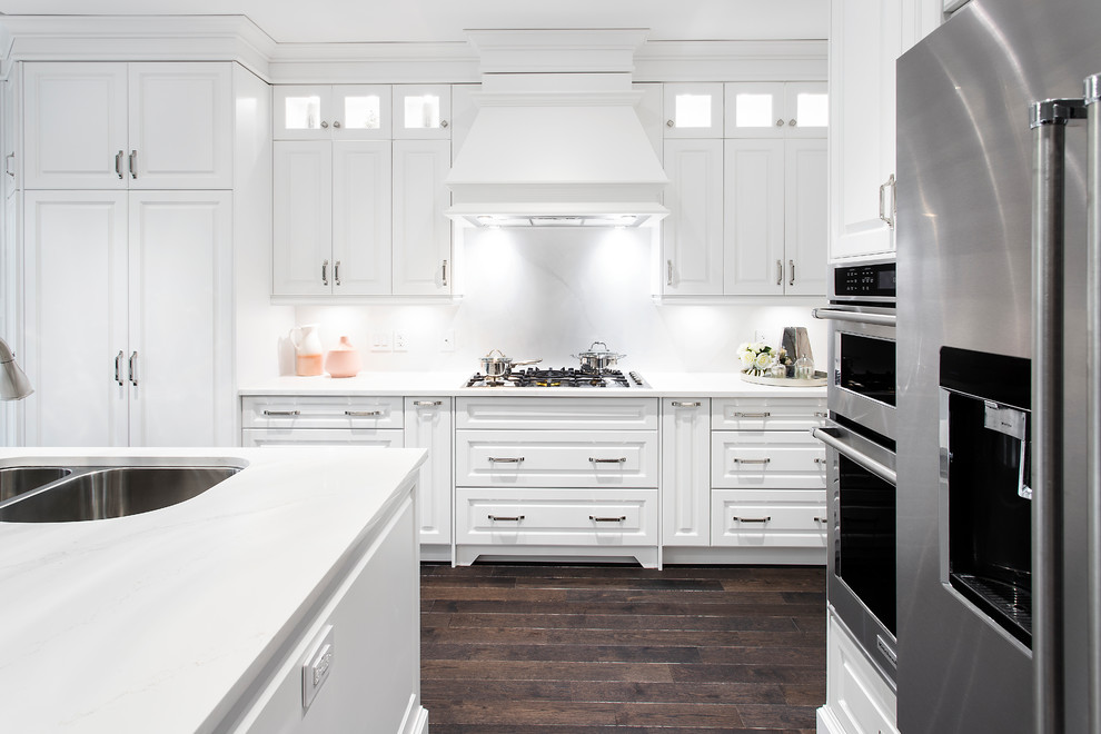 This is an example of a mid-sized transitional l-shaped kitchen in Toronto with an undermount sink, raised-panel cabinets, white cabinets, quartzite benchtops, white splashback, marble splashback, stainless steel appliances, dark hardwood floors, with island, brown floor and white benchtop.