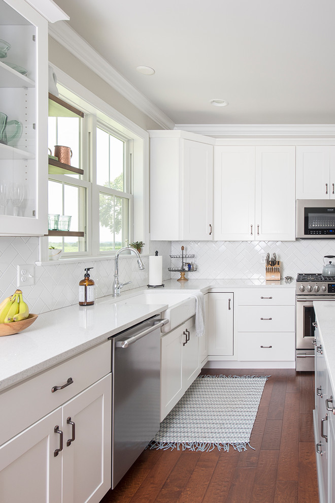 Example of a mid-sized transitional l-shaped medium tone wood floor and brown floor open concept kitchen design with a farmhouse sink, shaker cabinets, white cabinets, quartzite countertops, white backsplash, subway tile backsplash, stainless steel appliances and an island