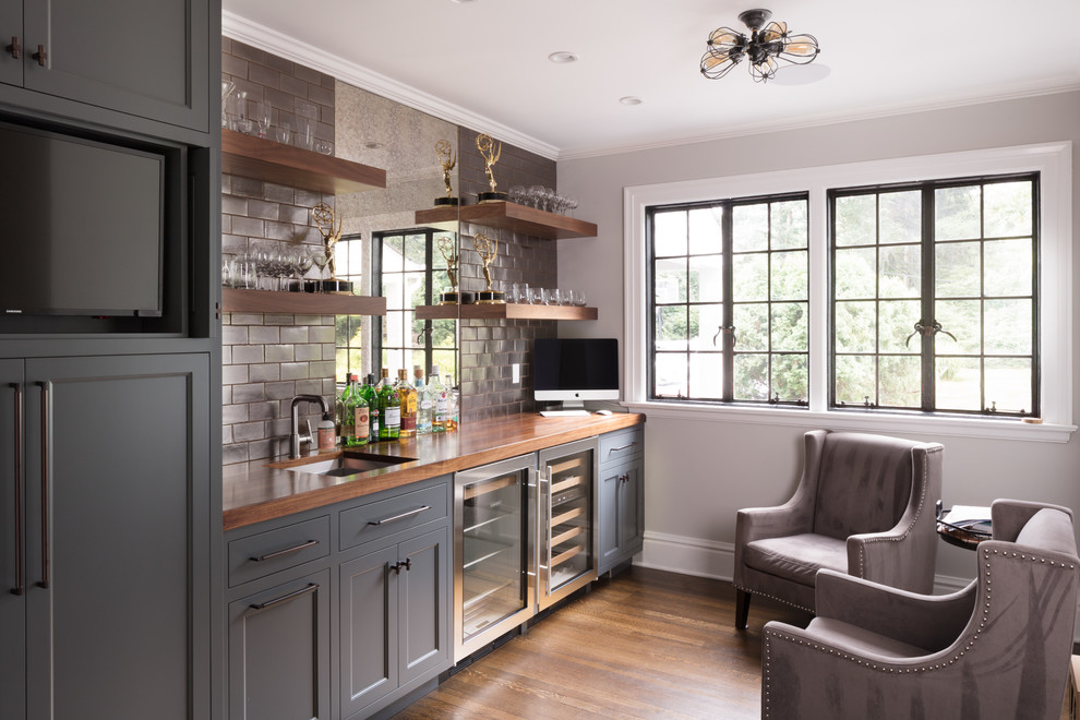 Photo of a traditional single-wall wet bar in New York with an undermount sink, recessed-panel cabinets, grey cabinets, wood benchtops, grey splashback, subway tile splashback, dark hardwood floors, brown floor and brown benchtop.