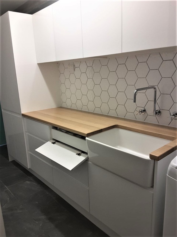 Inspiration for a mid-sized contemporary single-wall dedicated laundry room in Central Coast with a farmhouse sink, flat-panel cabinets, white cabinets, wood benchtops, white walls, ceramic floors, a stacked washer and dryer and black floor.