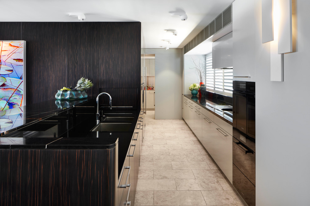 This is an example of a contemporary galley kitchen in Sydney with an undermount sink, flat-panel cabinets, dark wood cabinets, a peninsula, beige floor, black benchtop, marble benchtops, black splashback, stone slab splashback, black appliances and limestone floors.