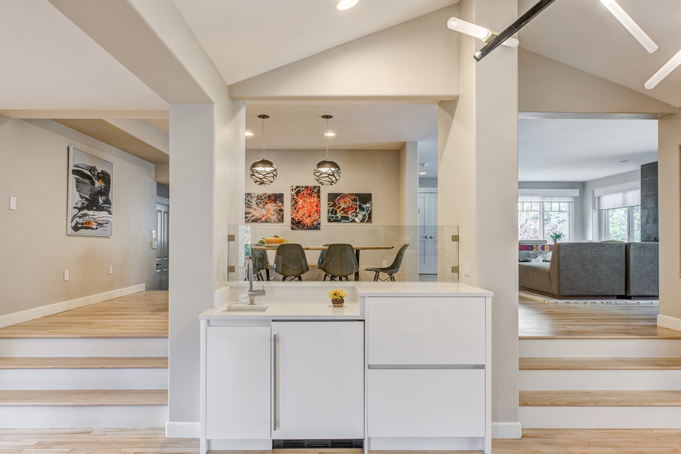 Photo of an expansive contemporary l-shaped home bar in Denver with an undermount sink, flat-panel cabinets, white cabinets, quartz benchtops, white splashback and light hardwood floors.