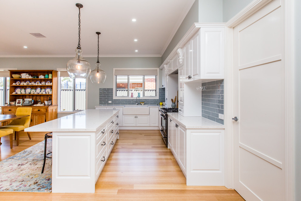 Photo of a mid-sized country l-shaped kitchen in Geelong with an integrated sink, raised-panel cabinets, white cabinets, grey splashback, ceramic splashback, medium hardwood floors, multiple islands and brown floor.