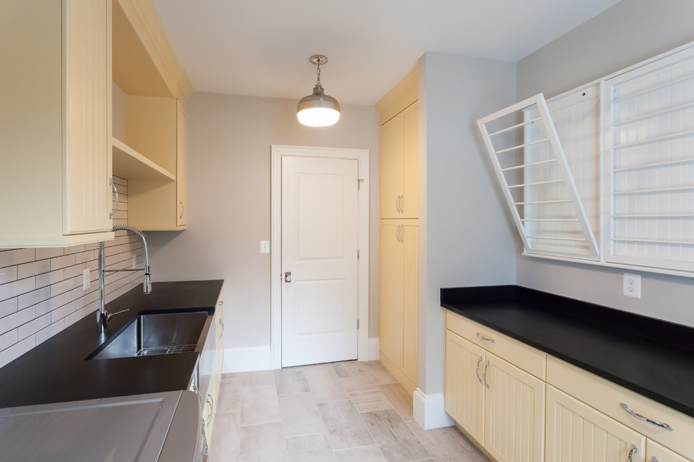 Photo of a country galley dedicated laundry room in DC Metro with a farmhouse sink, beaded inset cabinets, yellow cabinets, beige walls, a side-by-side washer and dryer, beige floor and black benchtop.