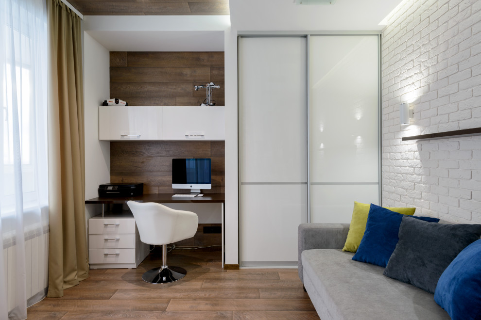 This is an example of a mid-sized contemporary home office in Novosibirsk with white walls, medium hardwood floors, a built-in desk, brown floor and brick walls.