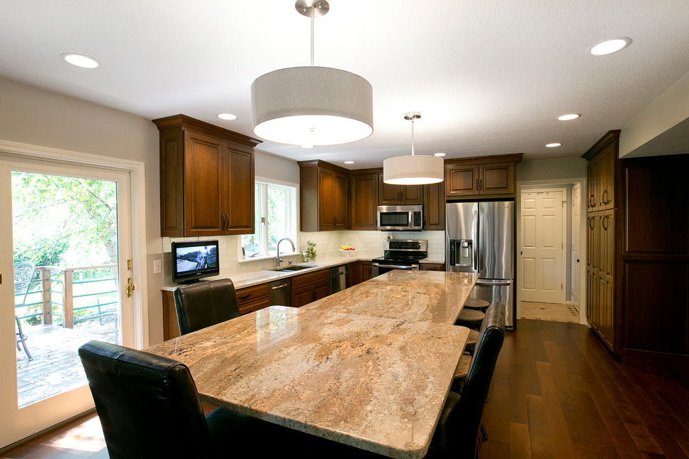 Inspiration for a transitional l-shaped medium tone wood floor eat-in kitchen remodel with a double-bowl sink, raised-panel cabinets, dark wood cabinets, granite countertops, white backsplash, glass tile backsplash, stainless steel appliances and an island