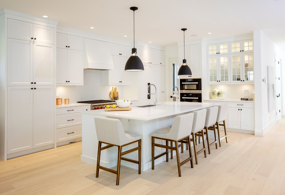 Inspiration for a country l-shaped kitchen in Vancouver with a double-bowl sink, shaker cabinets, white splashback, mosaic tile splashback, black appliances, light hardwood floors, with island, beige floor and white benchtop.