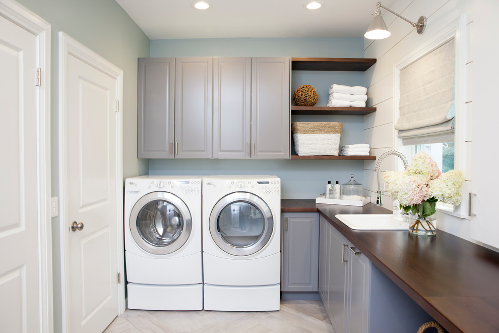 Design ideas for a large traditional l-shaped dedicated laundry room in New York with raised-panel cabinets, grey cabinets, wood benchtops, blue walls, porcelain floors, a side-by-side washer and dryer, a drop-in sink and brown benchtop.