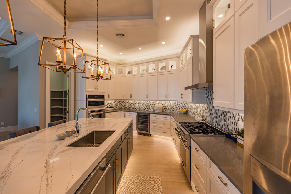 Large transitional l-shaped open plan kitchen in Miami with an undermount sink, shaker cabinets, white cabinets, marble benchtops, multi-coloured splashback, mosaic tile splashback, stainless steel appliances, light hardwood floors, with island and brown floor.