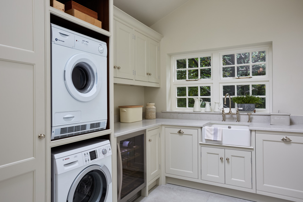 Design ideas for a mid-sized transitional l-shaped laundry room in London with a farmhouse sink, recessed-panel cabinets, grey cabinets, beige walls, porcelain floors, a stacked washer and dryer, grey floor and white benchtop.
