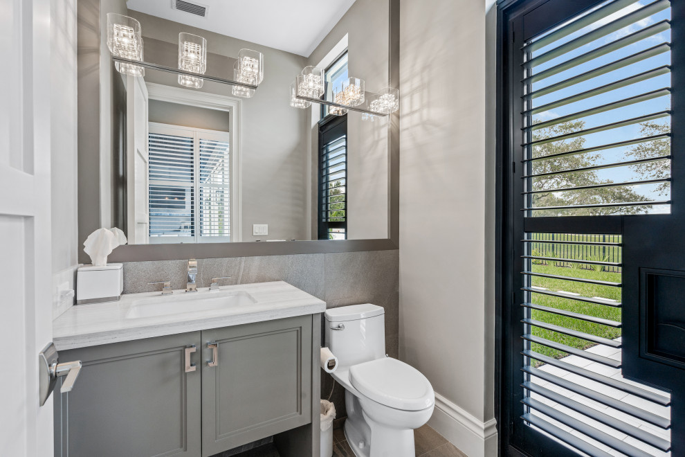 This is an example of a mid-sized modern powder room in Miami with recessed-panel cabinets, grey cabinets, a one-piece toilet, gray tile, porcelain tile, grey walls, porcelain floors, an undermount sink, marble benchtops, beige floor, white benchtops and a built-in vanity.