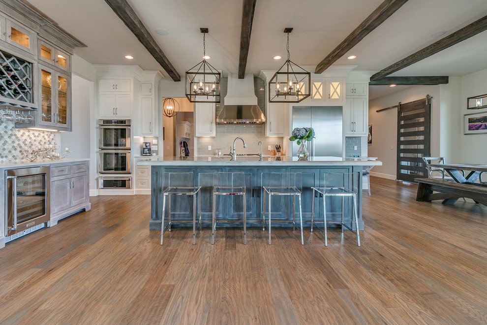This is an example of an expansive country l-shaped eat-in kitchen in Oklahoma City with a farmhouse sink, shaker cabinets, white cabinets, quartz benchtops, grey splashback, subway tile splashback, stainless steel appliances, light hardwood floors, with island and grey floor.