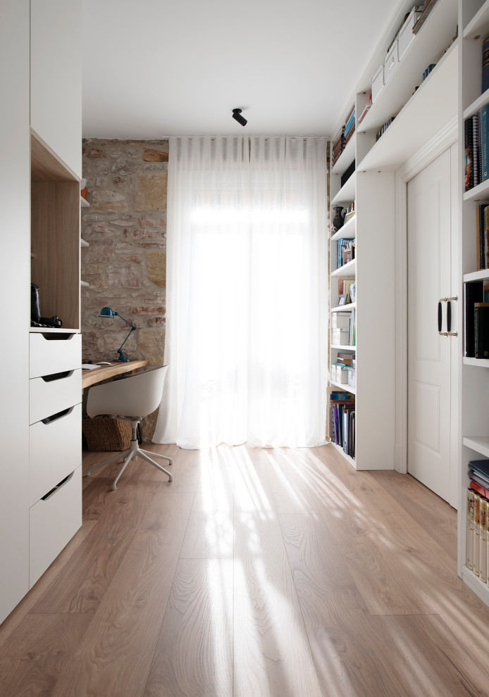 Cette image montre un bureau nordique de taille moyenne avec une bibliothèque ou un coin lecture, un mur blanc, sol en stratifié, un bureau intégré, un sol marron et poutres apparentes.