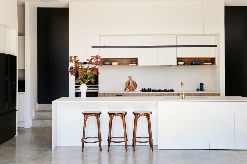 Photo of a large contemporary l-shaped kitchen in Geelong with an undermount sink, flat-panel cabinets, white cabinets, black appliances, with island, grey floor, white benchtop, white splashback and concrete floors.