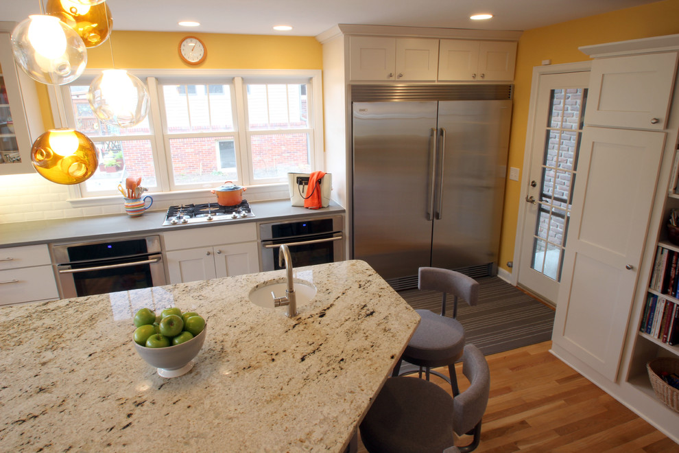 Eat-in kitchen - large transitional l-shaped light wood floor eat-in kitchen idea with a farmhouse sink, shaker cabinets, white cabinets, glass countertops, white backsplash, subway tile backsplash, stainless steel appliances and an island
