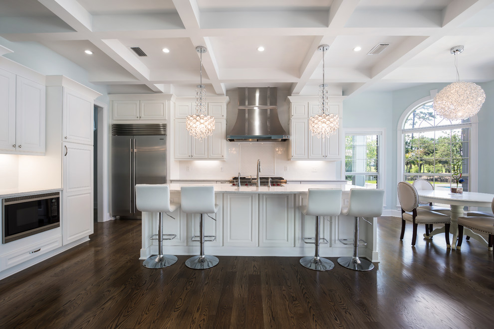 This is an example of a mid-sized contemporary l-shaped eat-in kitchen in Jacksonville with a farmhouse sink, raised-panel cabinets, white cabinets, quartz benchtops, white splashback, subway tile splashback, stainless steel appliances, dark hardwood floors, with island, brown floor and white benchtop.
