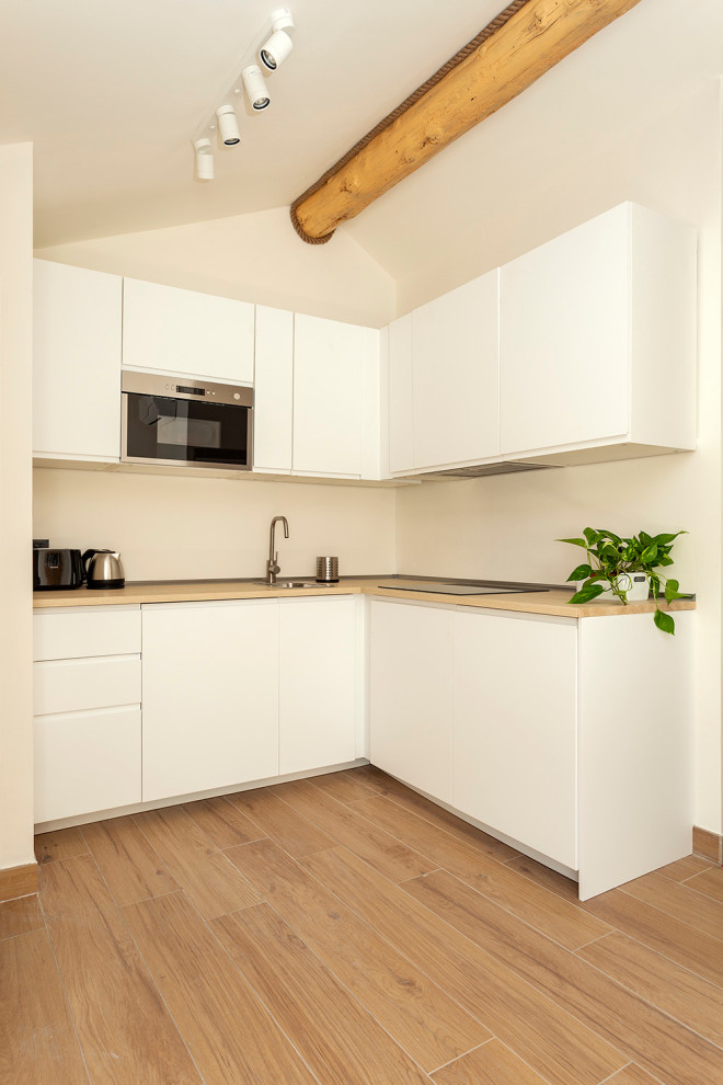 Idée de décoration pour une cuisine américaine minimaliste en L avec un placard à porte plane, des portes de placard blanches, un plan de travail en bois, parquet clair et poutres apparentes.